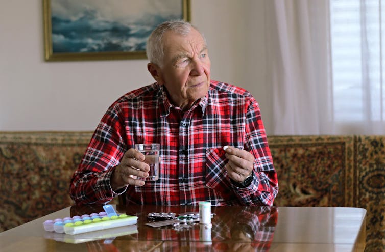 A senior man taking a tablet. There are a variety of medications on the table.