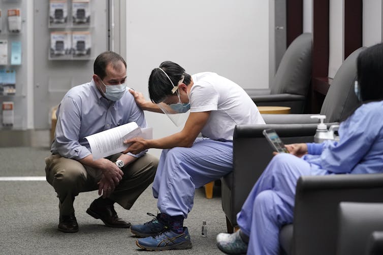 A man in scrubs and a face shield reaches out to a man in a face mask.