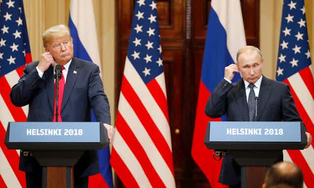 US president Donald Trump (right) and Vladimir Putin at a press conference in Helsinki in 2018. They are both standing at podiums and adjusting their earpieces with Russian and US flags inthe background.