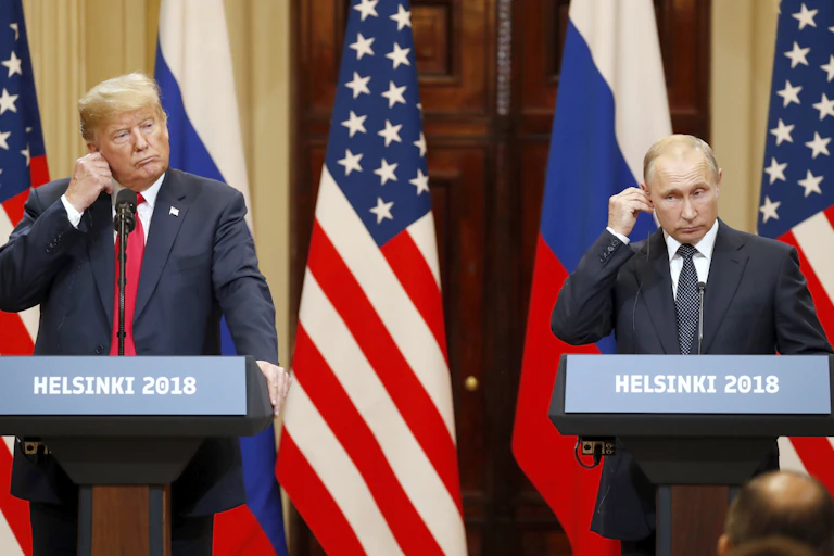 US president Donald Trump (right) and Vladimir Putin at a press conference in Helsinki in 2018. They are both standing at podiums and adjusting their earpieces with Russian and US flags inthe background.