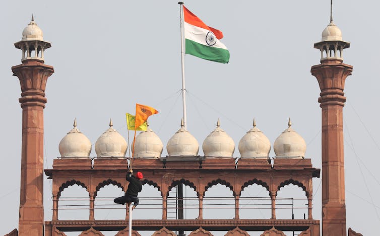 A man climbs a flag pole to raise a yellow and orange flag in front on Delhi's Red Fort and the Indian national flag
