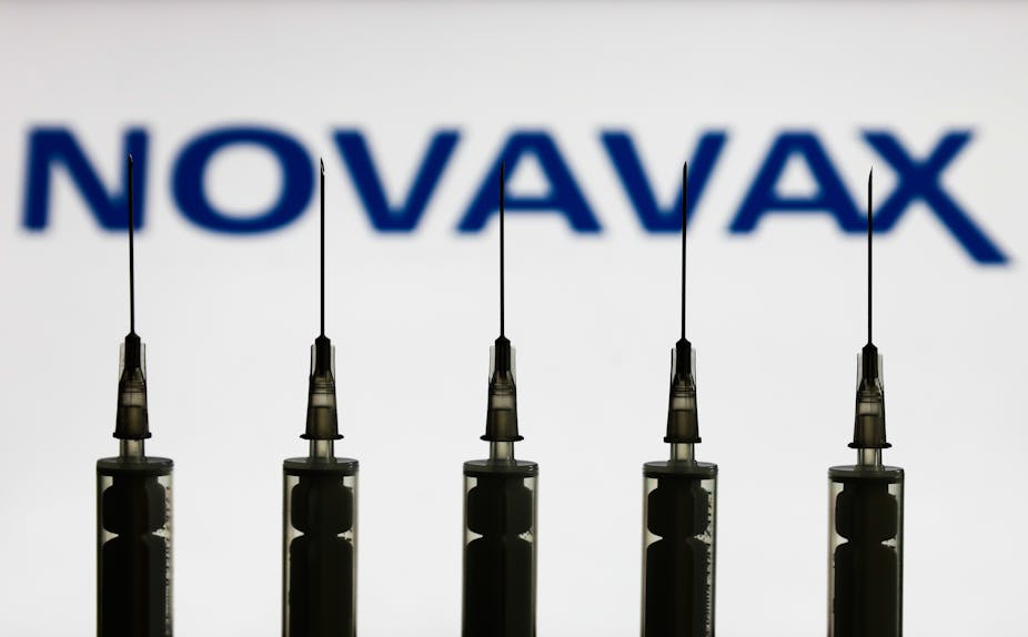 Medical syringes are seen with Novavax company logo displayed on a screen in the background
