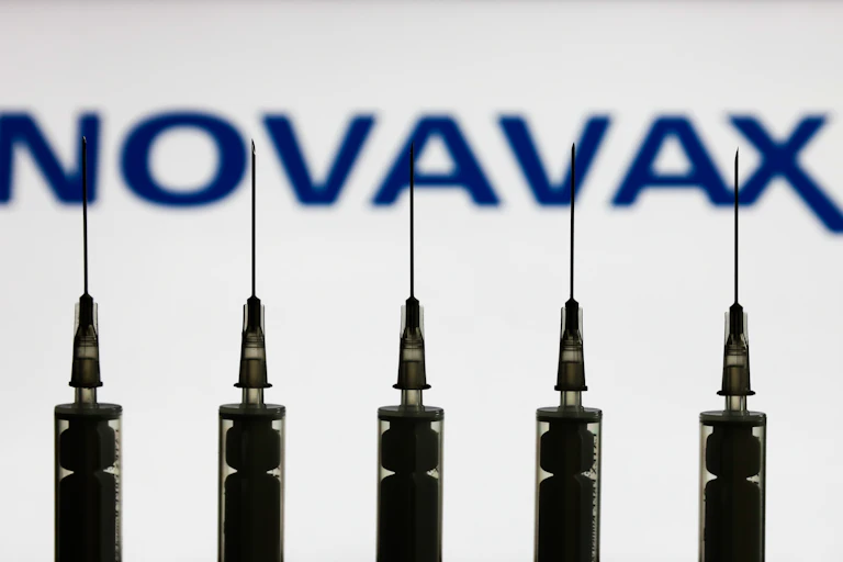 Medical syringes are seen with Novavax company logo displayed on a screen in the background