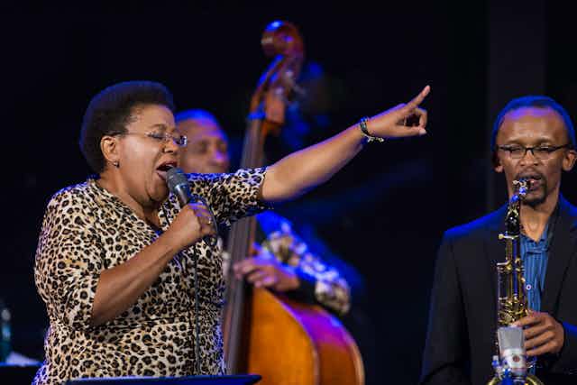 A woman in a leopard print dress and short Afro hairstyle closes her eyes as she sings into a microphone, her finger pointing in the air ahead of her. Around her are jazz musicians playing live.