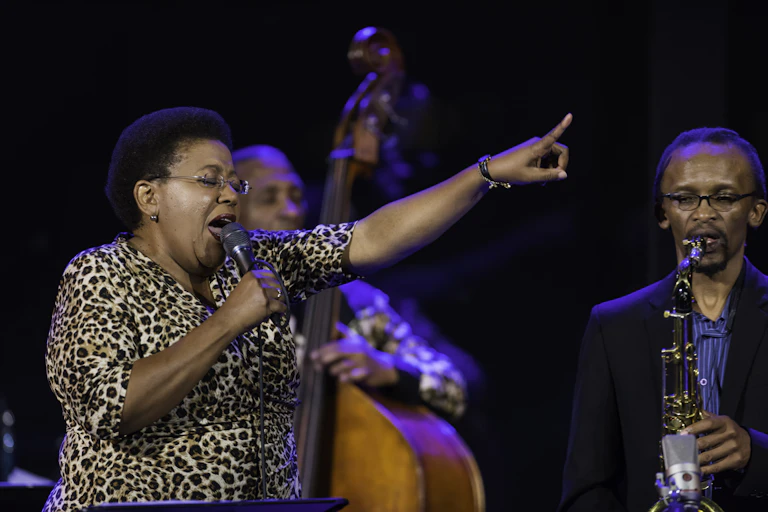 A woman in a leopard print dress and short Afro hairstyle closes her eyes as she sings into a microphone, her finger pointing in the air ahead of her. Around her are jazz musicians playing live.