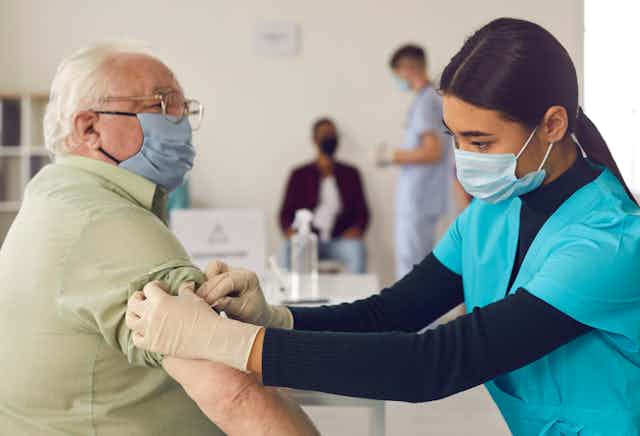 A nurse in a mask administers a COVID vaccine to an elderly man in a mask.