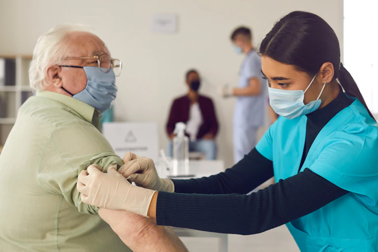 A nurse in a mask administers a COVID vaccine to an elderly man in a mask.