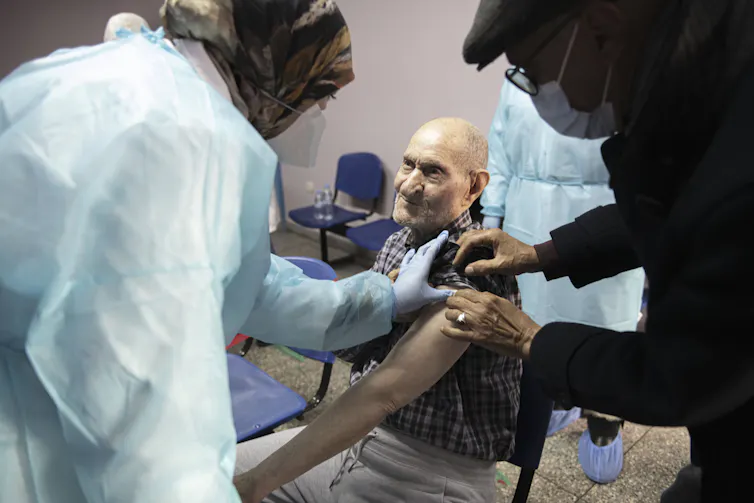 A man in a waiting room helped by two others to roll up his sleeve