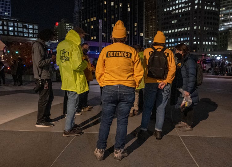 People in orange and yellow sweatshirts labeled 'Defenders' talk in a circle at night