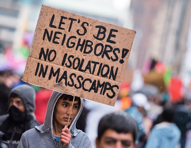 Demonstrator holding a sign reading 'Let's be neighbors. No isolation in masscamps'