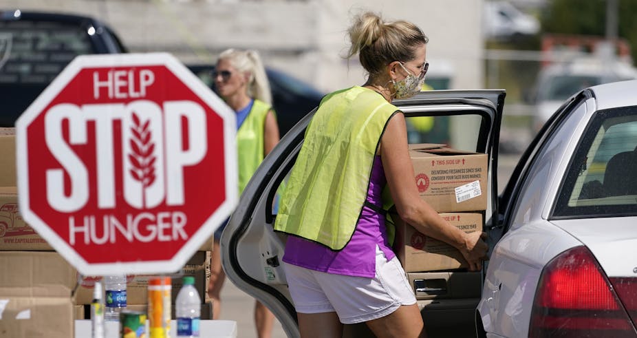 A woman in shorts and a yellow vest loads boxes into a car while standing next to a sign that says 'Help Stop Hunger.'