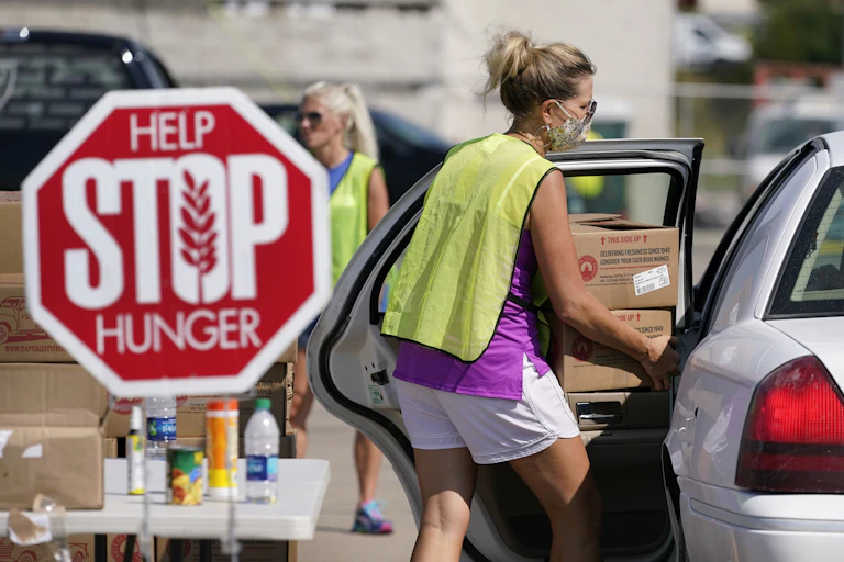 A woman in shorts and a yellow vest loads boxes into a car while standing next to a sign that says 'Help Stop Hunger.'