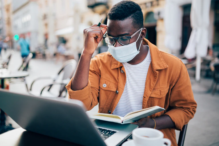 A Black man sits down and reads a book while his computer is open on a desk.