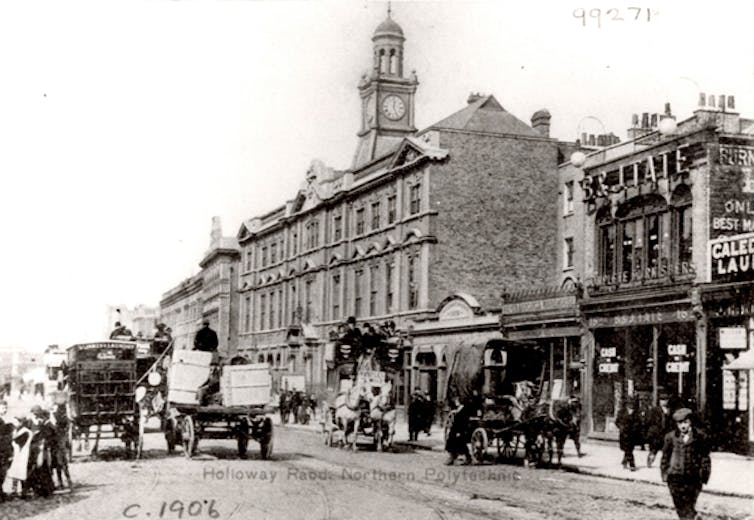 Black and white photo of large building with clock tower, with horse-drawn carts and carriages passing in front.