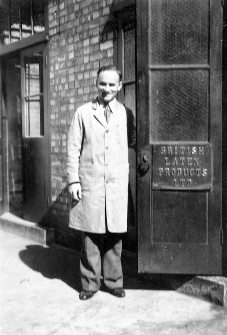 Black and white photo of smiling man in warehouse coat stood outside door with sign for 'British Latex Products'.