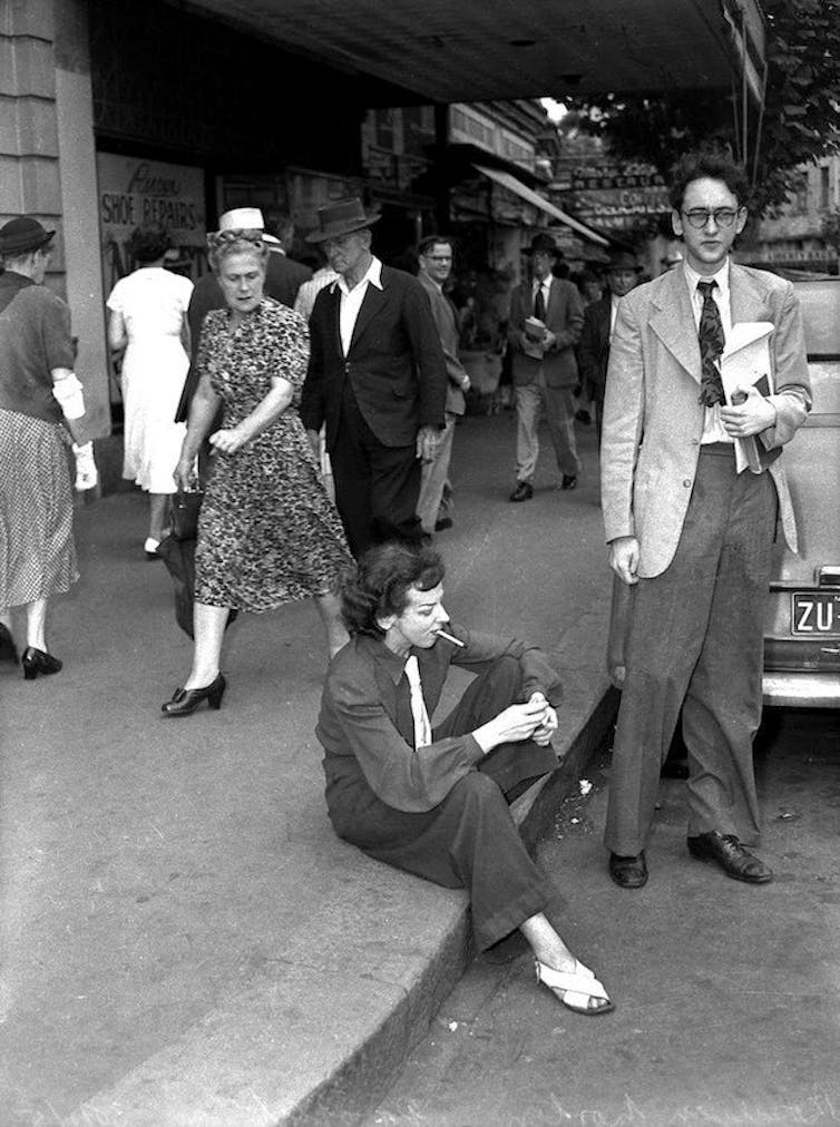 Norton wearing slacks, smoking in public, sitting in the gutter. A woman walks past, looking scandalised.