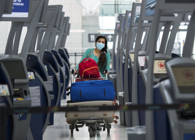 A woman wearing a face mask and pushing a full luggage cart walks between Air Canada kiosks