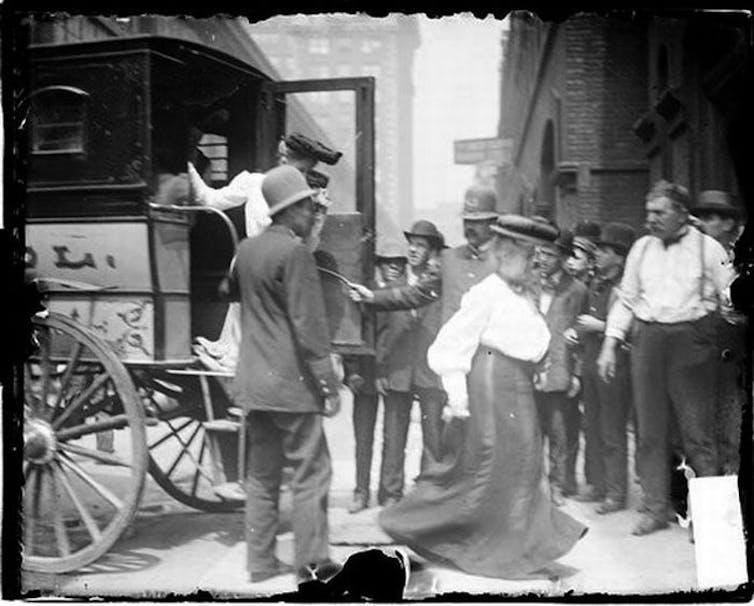 Image of two women exiting a police van past two policemen and a crowd of people watching on the sidewalk.