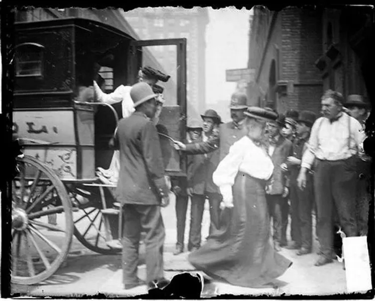 Image of two women exiting a police van past two policemen and a crowd of people watching on the sidewalk.