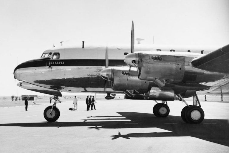 An 1950s aeroplane stands on the tarmac with British colonial servicemen in the background.