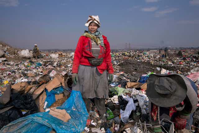A woman dressed in layers of dirty clothing looks into the camera in a sea of rubbish at a dump.