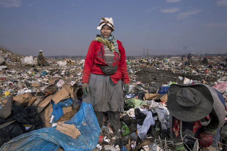 A woman dressed in layers of dirty clothing looks into the camera in a sea of rubbish at a dump.