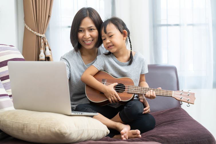 Image of a mother and daughter using laptop studying to play ukulele at home.