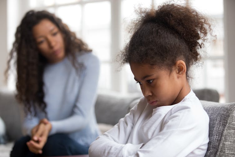 A child folds her arms in a grumpy posture while her mother looks on concerned