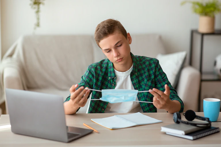 A boy holds a protective face mask while sitting at a table.