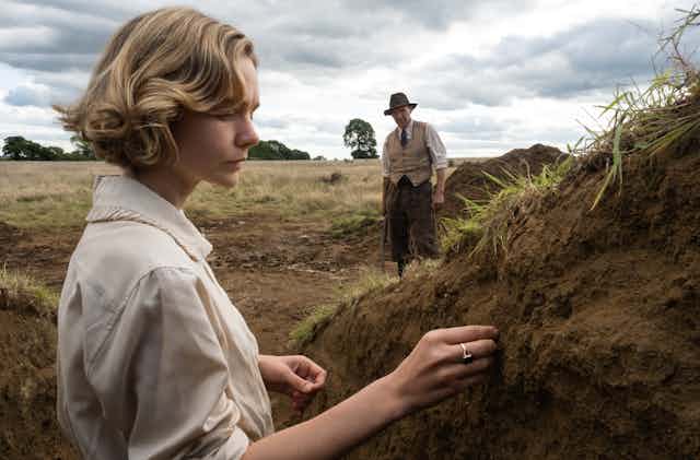 Carey Mulligan examines soil in an excavation pit  with Ralph Fiennes in background.