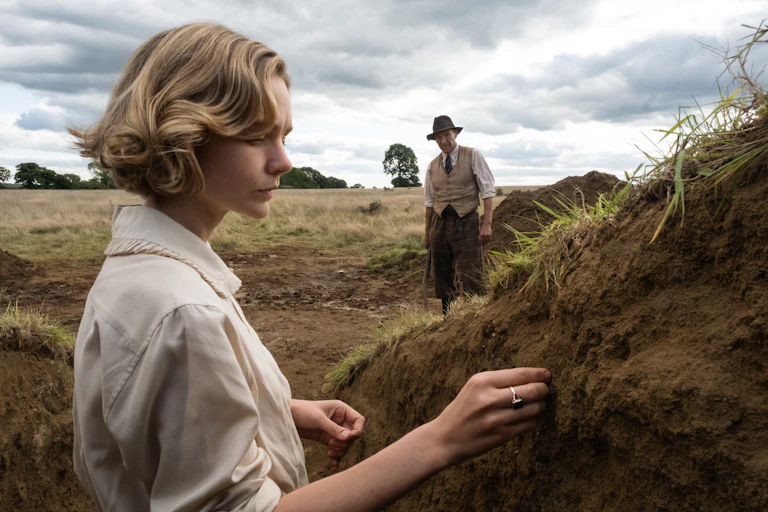 Carey Mulligan examines soil in an excavation pit with Ralph Fiennes in background.