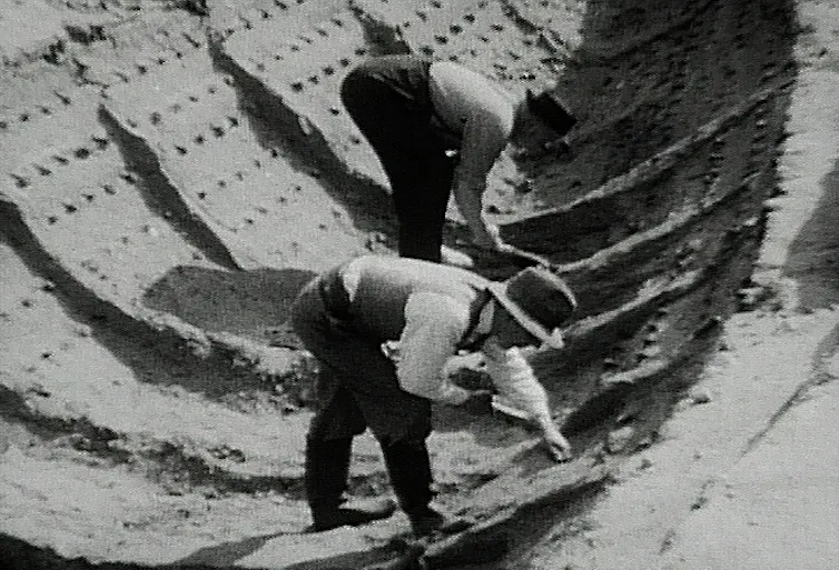 Black and white photo of two men bending over to study ship imprint in the soil.