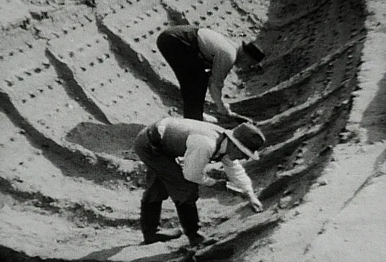 Black and white photo of two men bending over to study ship imprint in the soil.