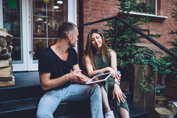 A couple sit on the doorstep talking. The man is holding a tablet computer.