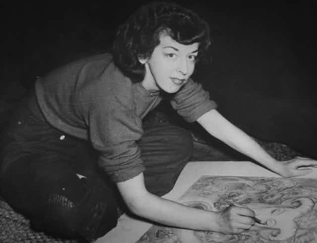 Black and white photo, a woman looks to camera while drawing.