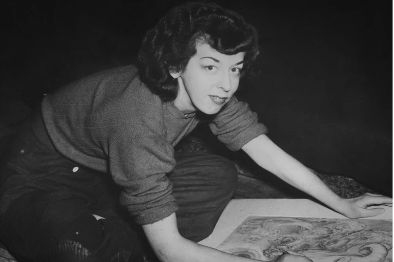 Black and white photo, a woman looks to camera while drawing.