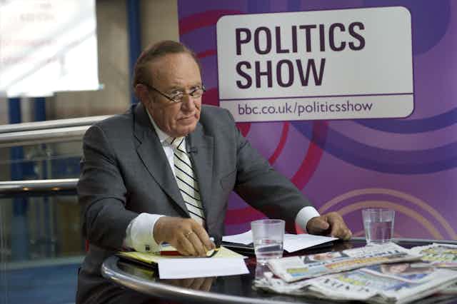 BBC politics broadcaster Andrew Neil sitting at a desk covered in newspapers in front of a sign saying: Politics Show.