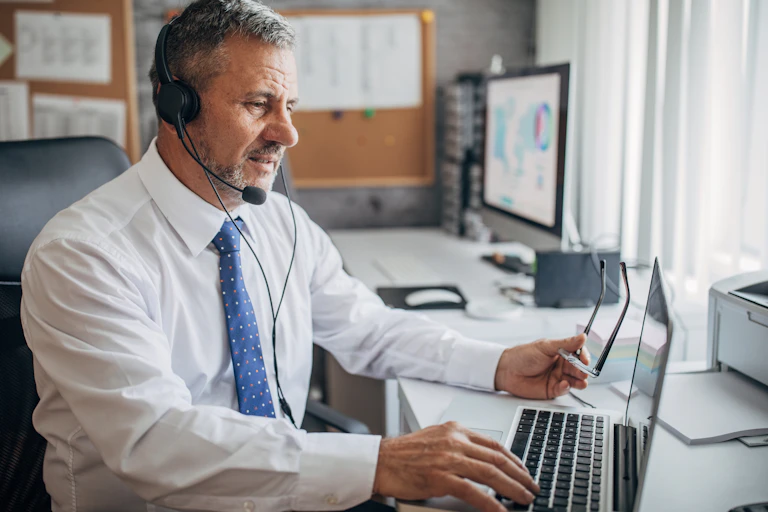 A male educator sits at a laptop wearing headphones and holding his eyeglasses.