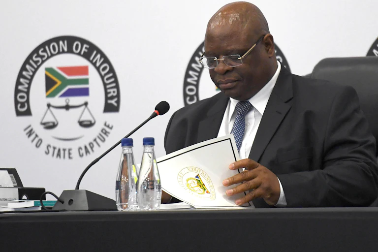 A bald-headed man wearing spectacles, a dark suit, white shirt and blue tie reads from a document