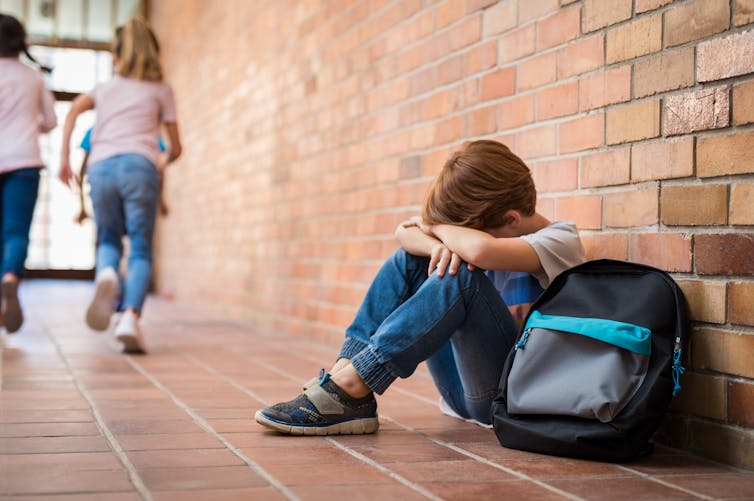 Image of boy sitting alone on floor after suffering an act of bullying while children run in the background.