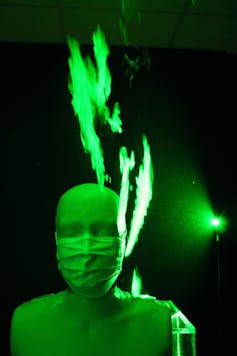 A mannequin wearing a mask lit by green light revealing cloud of vapour leaking from mask.