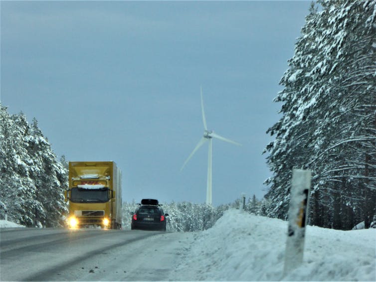 A snowy road traversed by a lorry and van with a wind turbine in the background.