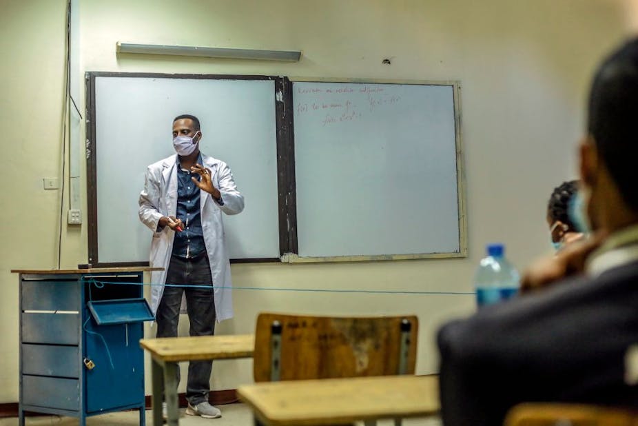 A teacher in Ethiopia wears a face mask and stands behind a blue thread-line denoting a boundary between him and the students.