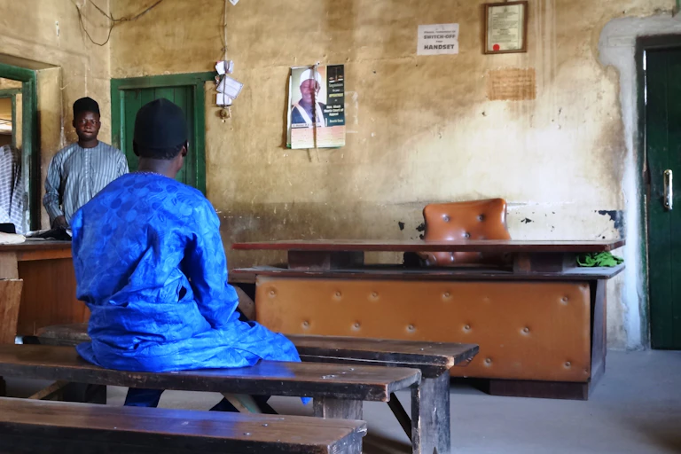 Man wearing a blue caftan and black cap sitting on a stool in a building