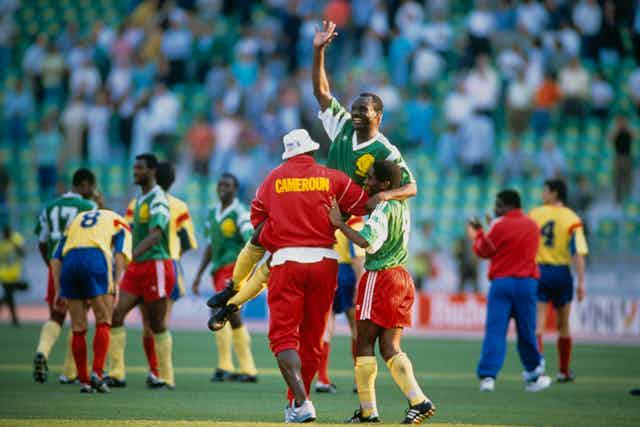A smiling man in football gear raises a hand in the air as he is held aloft by two other players, one in a red tracksuit with the word Cameroun in yellow, other players and arrows of spectators in the background.