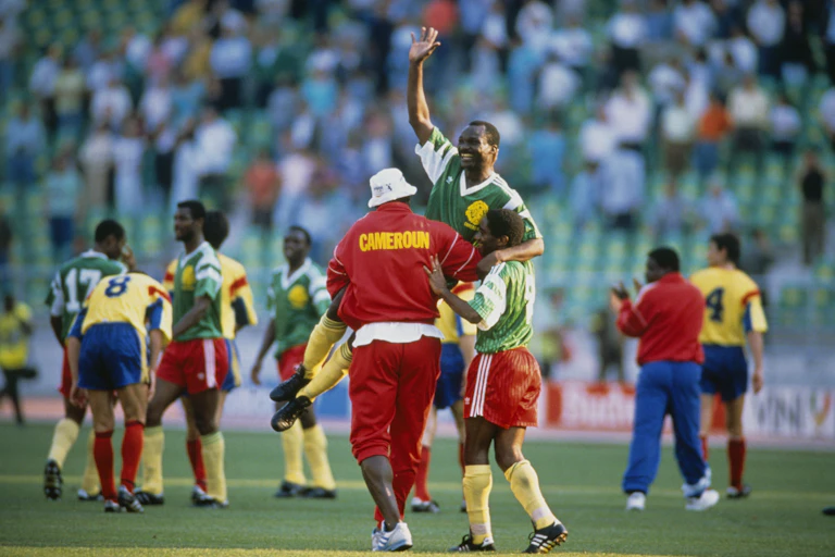 A smiling man in football gear raises a hand in the air as he is held aloft by two other players, one in a red tracksuit with the word Cameroun in yellow, other players and arrows of spectators in the background.