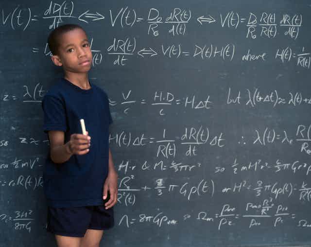 A Black boy holds a piece of chalk while standing by a chalk board filled with math equations.