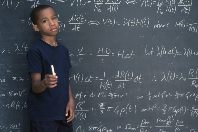 A Black boy holds a piece of chalk while standing by a chalk board filled with math equations.
