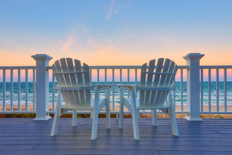 Two empty white adirondack chairs facing the ocean