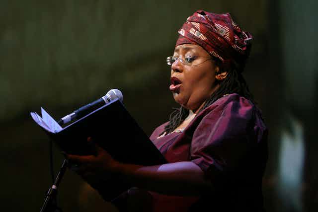 A woman dressed in purple, her hair covered by a traditional cloth and wearing glasses, sings into a mic as she reads music that she holds.